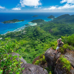man hiker tourist standing high above indian ocean on granit rock of tropical island  mountains taking pictures